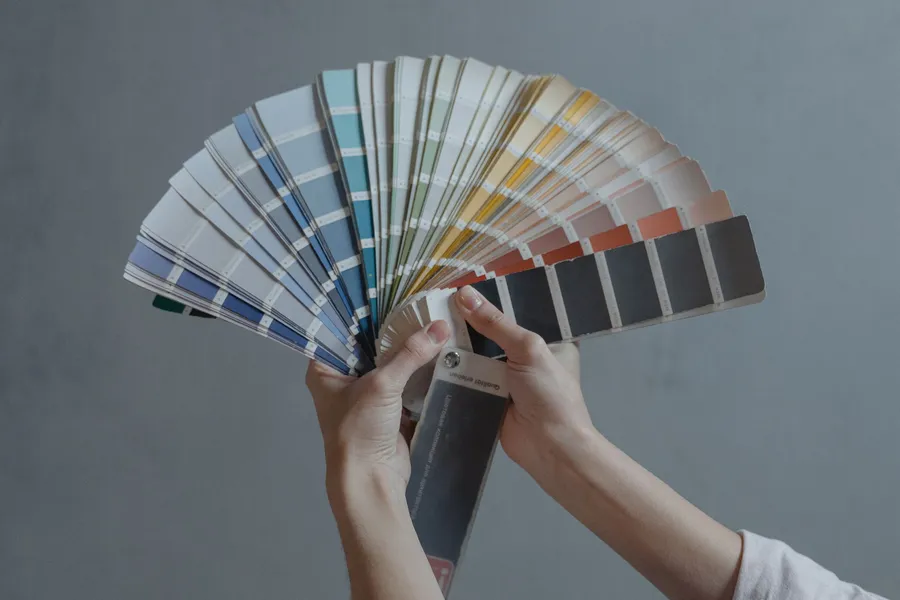 A hand holding an open fan deck of paint color samples against a painted wall.