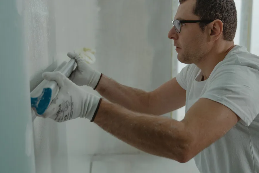 A worker in gloves smoothing filling compound across a repaired patch on an interior wall.
