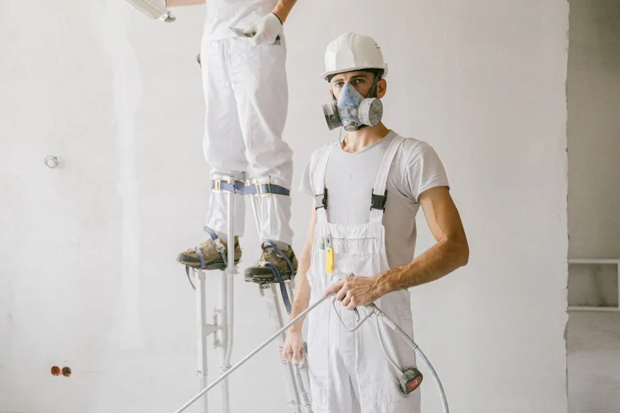 Two painters in coveralls, respirators and hard hats working on stilts to reach a high interior wall.