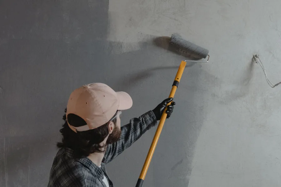 A painter using an extension-pole roller to lay a smooth coat of gray paint across an interior wall.