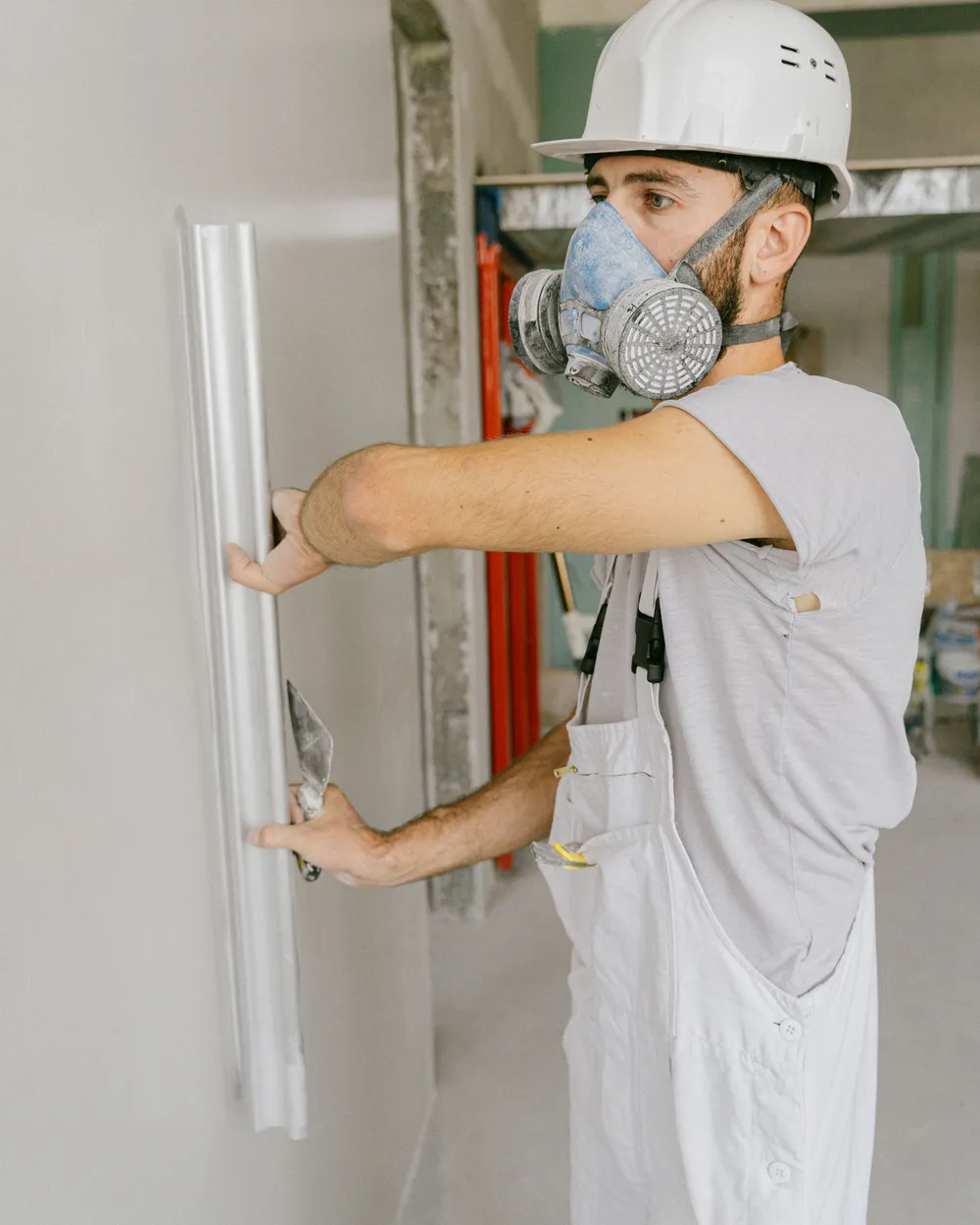 A worker in coveralls, a respirator and a hard hat holding a straight edge against a repaired white wall.