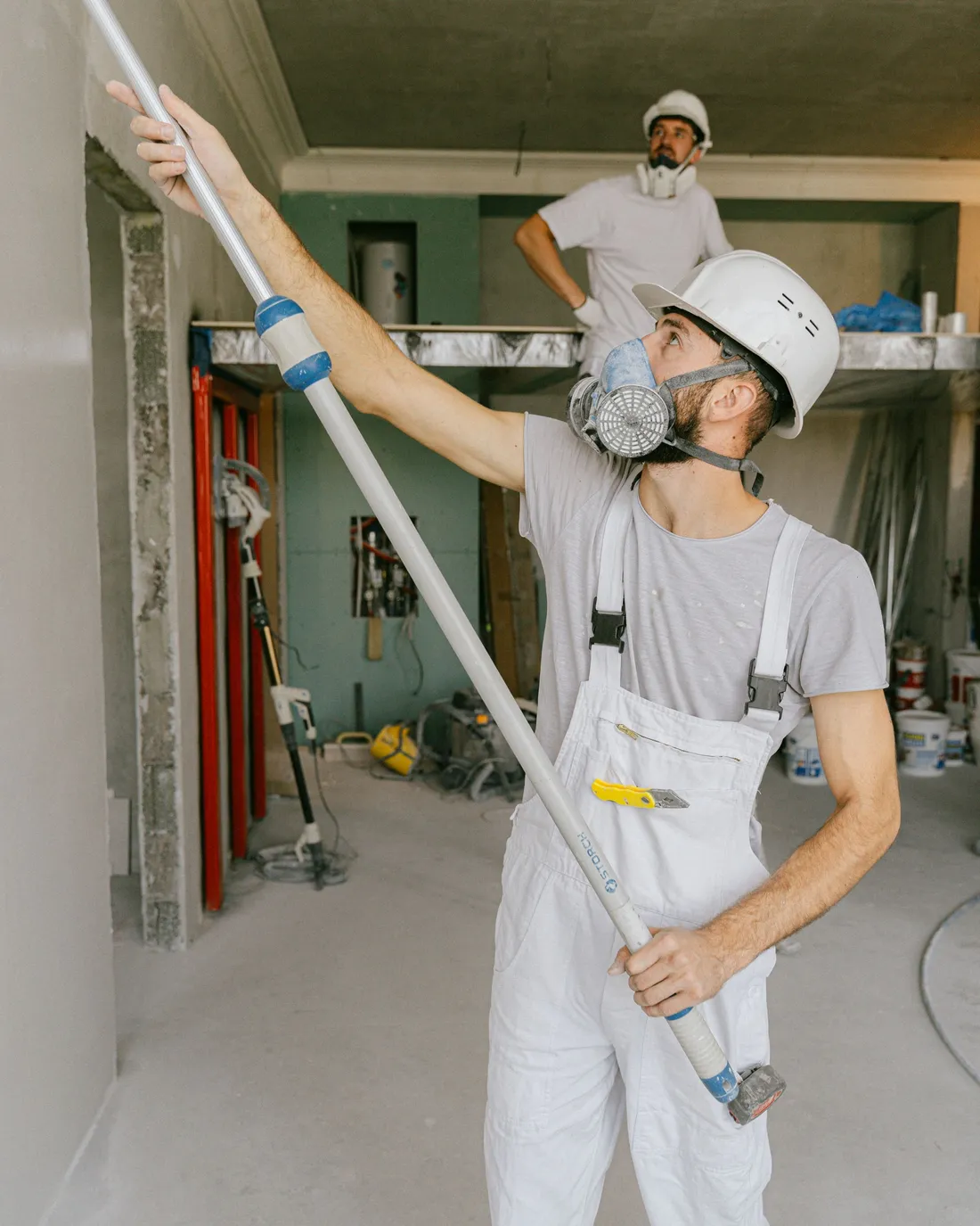 Two painters in white coveralls and protective masks coating an interior wall, one working with an extension-pole roller.