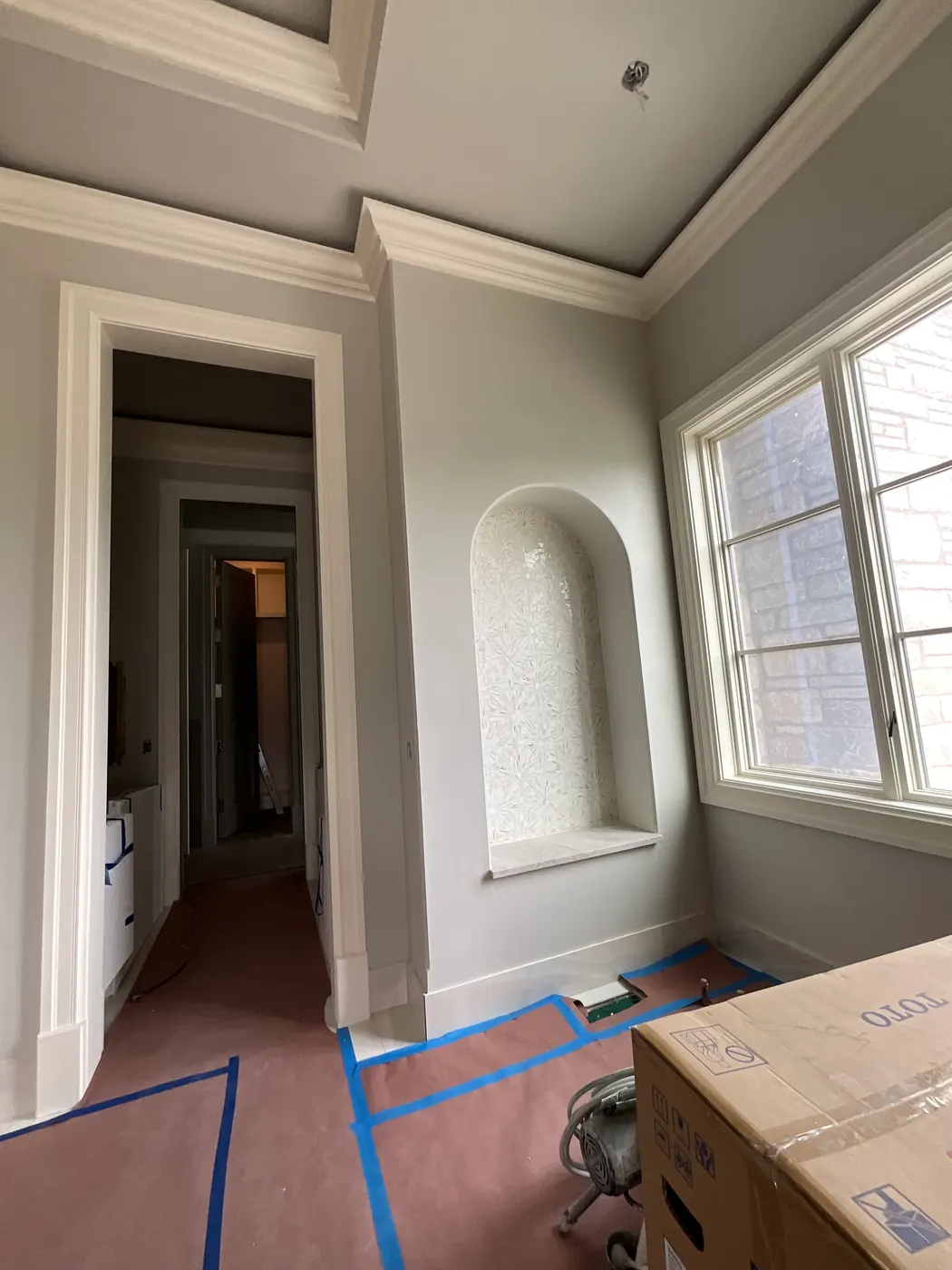 A room finished in soft gray-green with white crown molding, an arched niche tiled in white and a tall window in painted trim, the floor covered with taped protective paper.