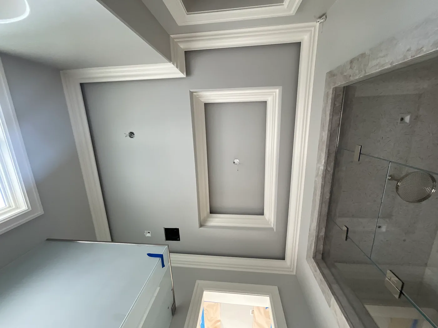 A bathroom with gray walls and white trim, including a large recessed molding panel, crown molding and a tiled shower behind glass, with the vanity masked off in plastic and tape.