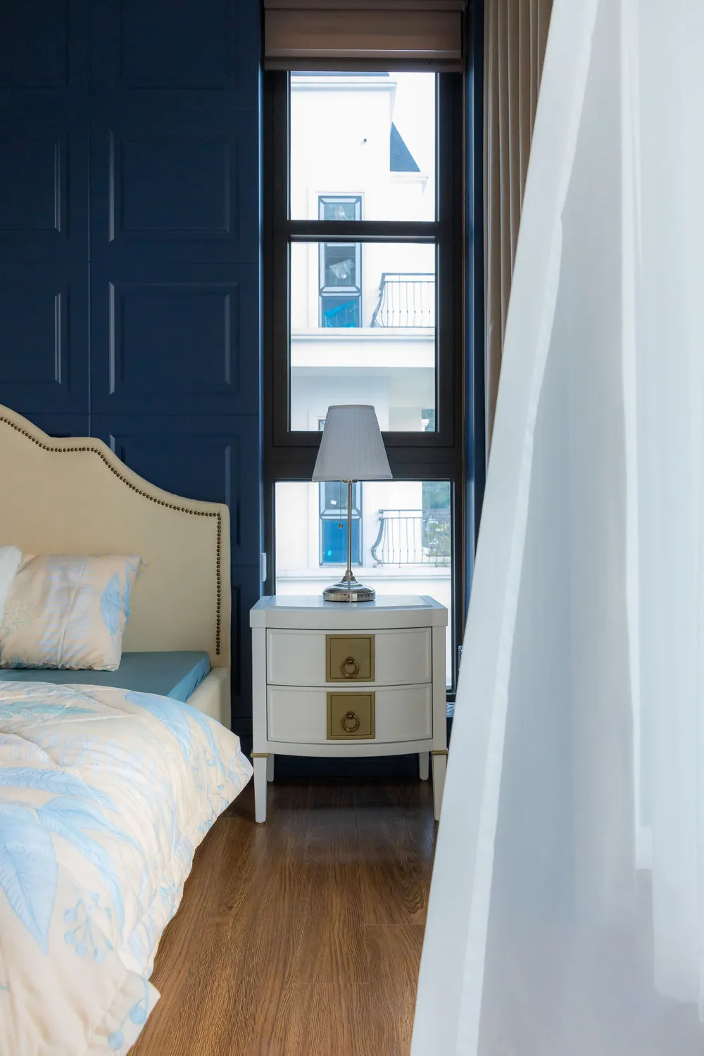A bedroom with a navy blue accent wall behind an upholstered headboard, white trim and a bright window.