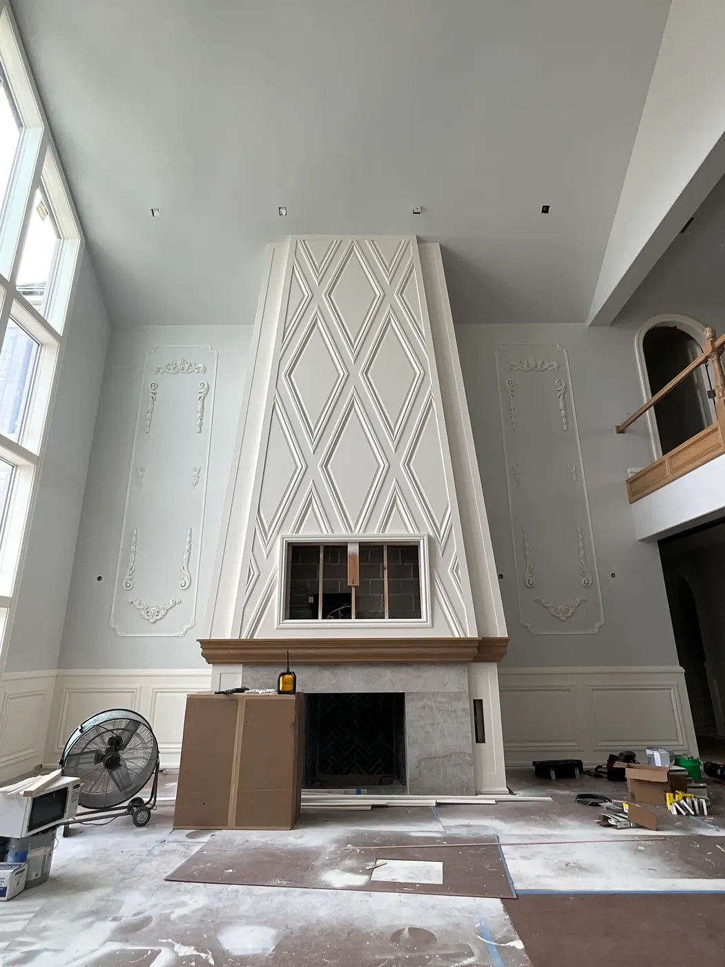A two-story room with a tapered white chimney breast finished in a diamond lattice of applied molding, flanked by ornamental wall panels above a wood-toned mantel and stone surround, with the floor covered against paint.