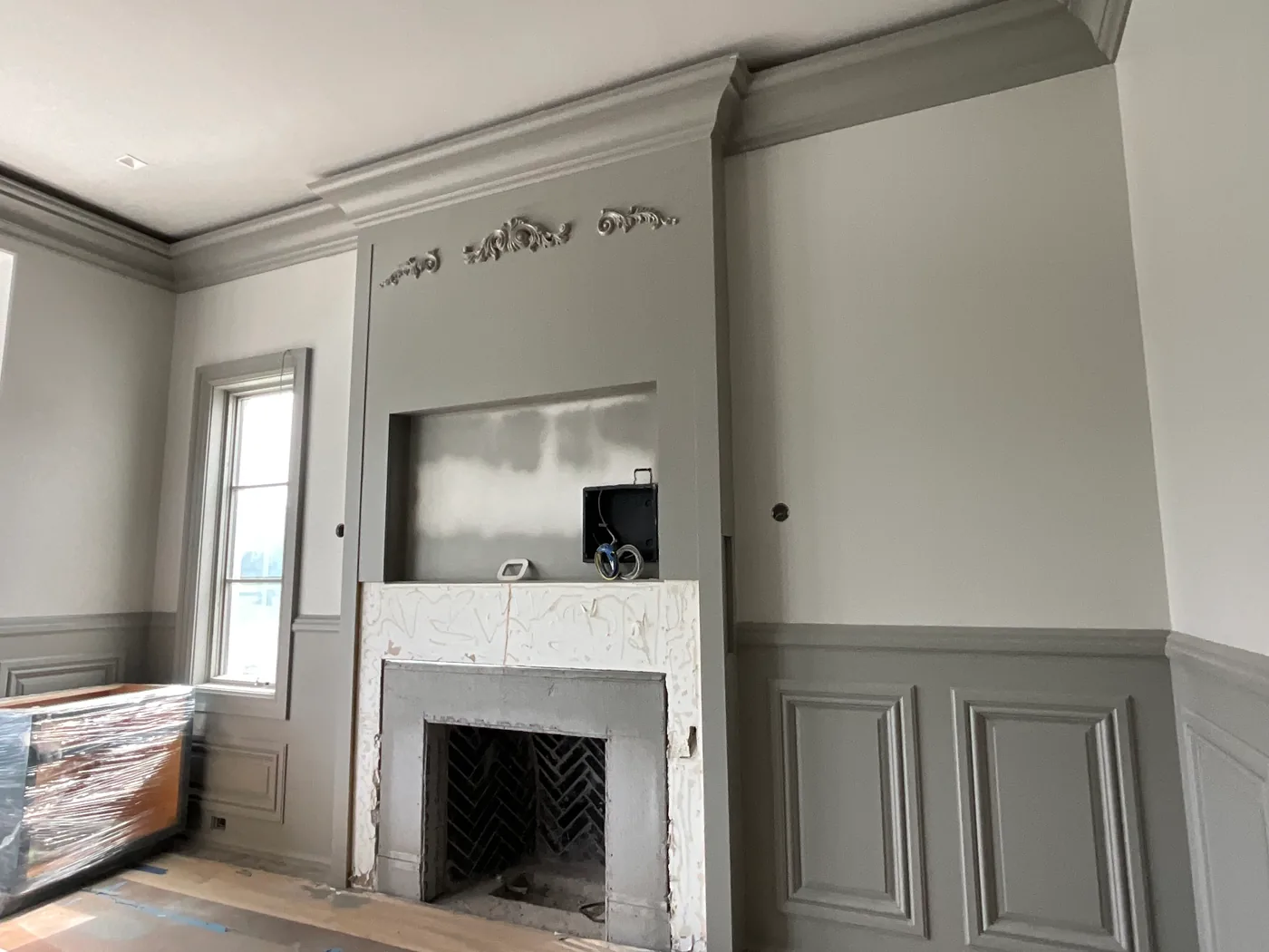 A fireplace wall painted warm gray, with crown molding, raised-panel wainscoting, a carved applique above the firebox and drop-covered floors.