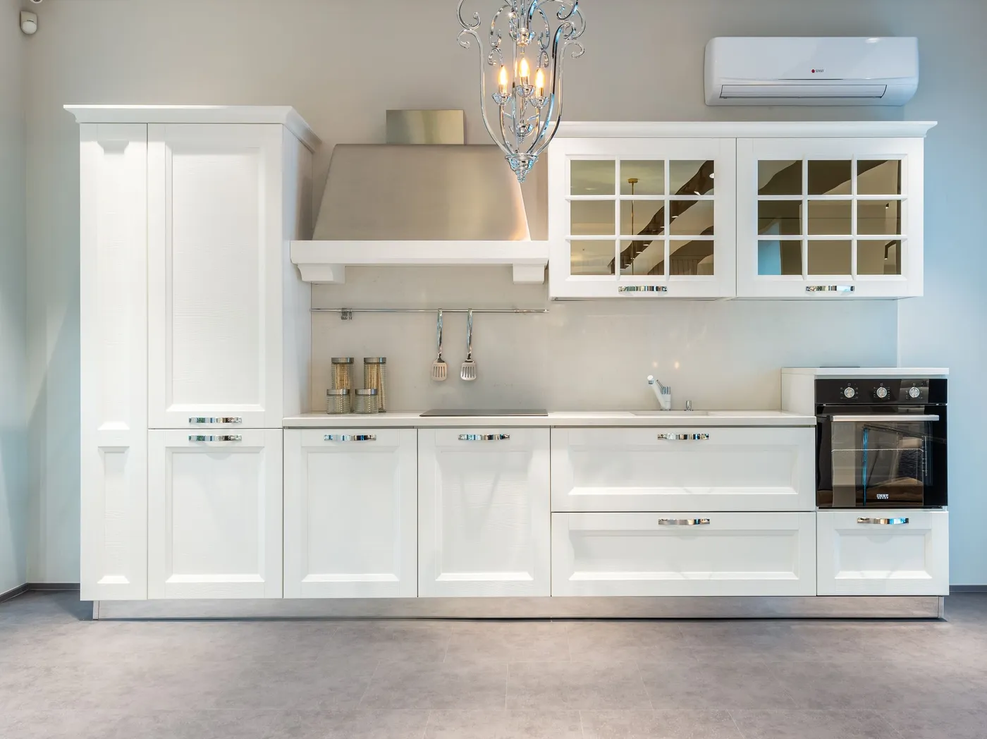 A kitchen with white shaker-style cabinets, glass upper doors and a chandelier above the run of units.