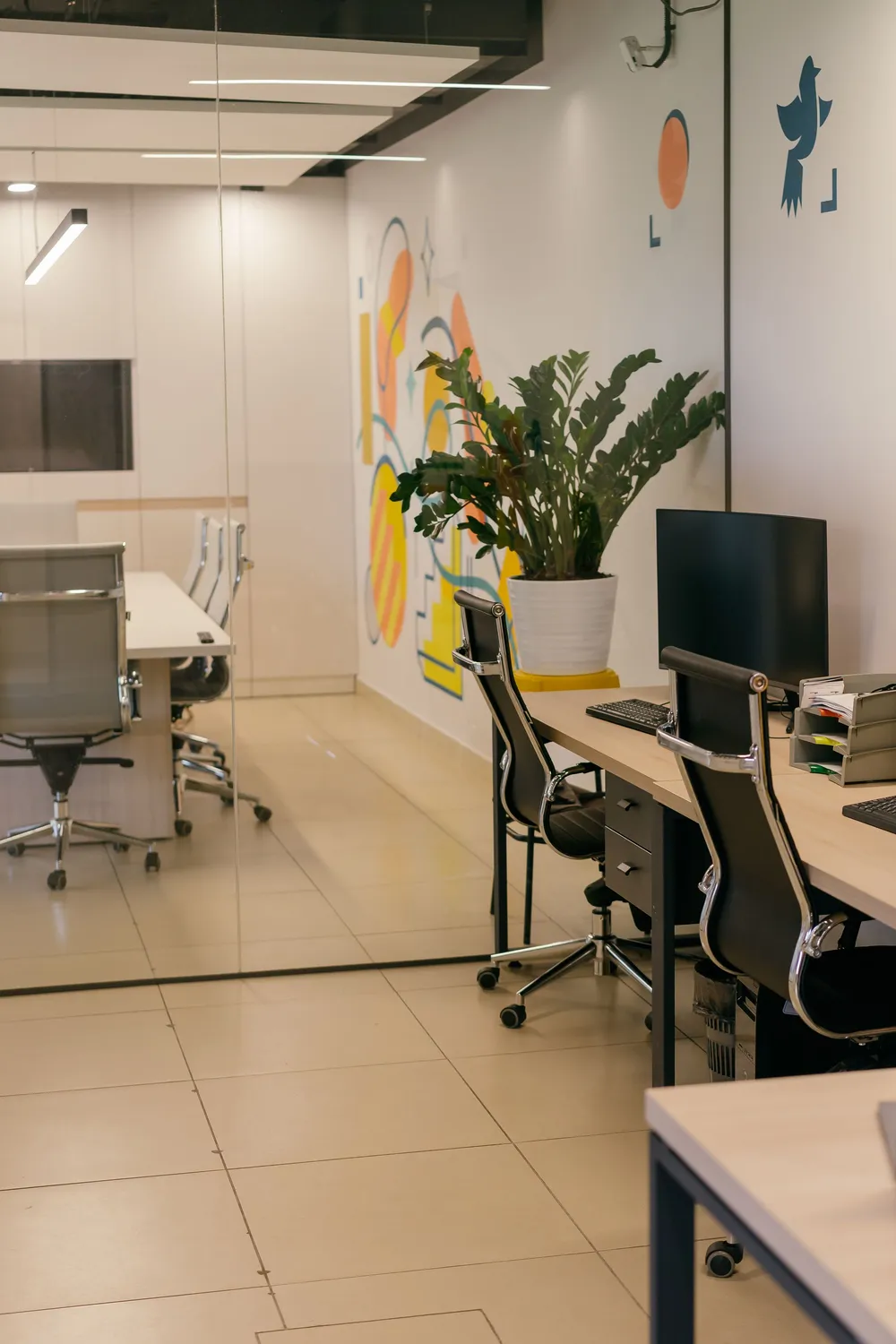 A bright office workspace with painted walls, a mural-style artwork, desks, task chairs and a large plant.