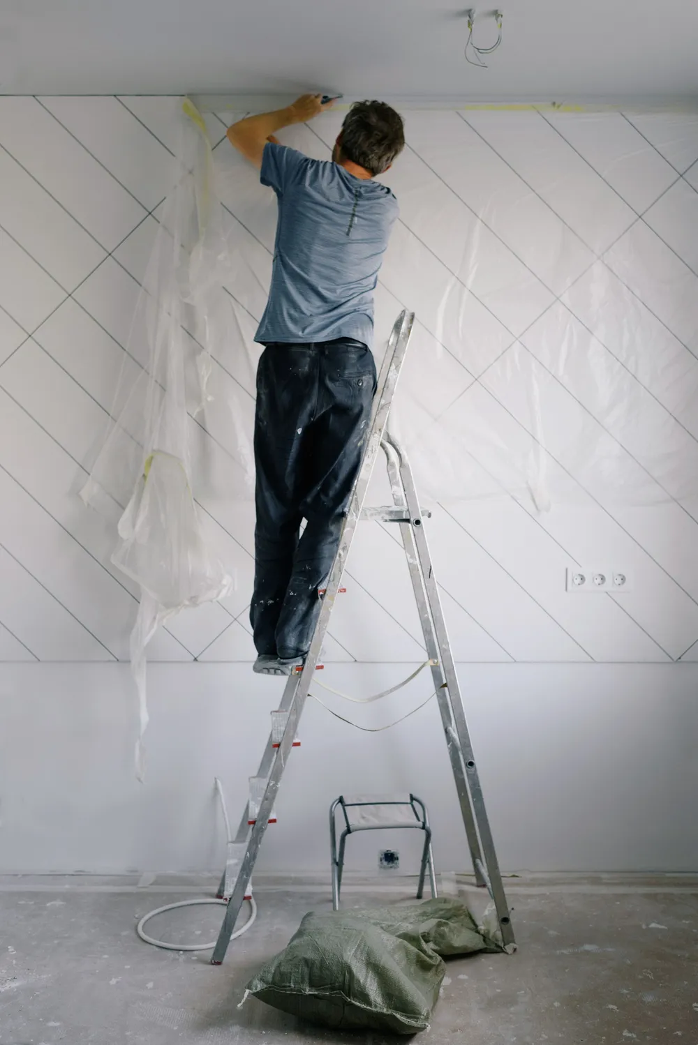 A painter on a ladder rolling white paint onto a tall interior wall, with the floor fully covered by drop cloths.