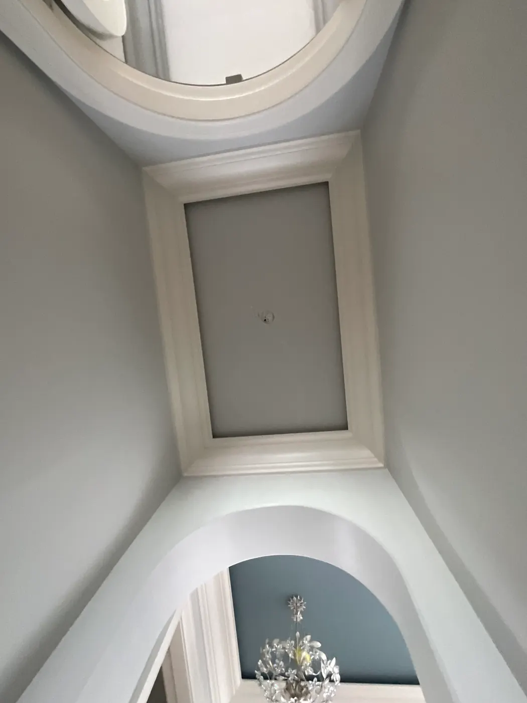 Looking up a stairwell to a recessed ceiling panel framed in wide white molding, with a curved opening above and, through the arch below, a crystal chandelier against a pale blue ceiling.
