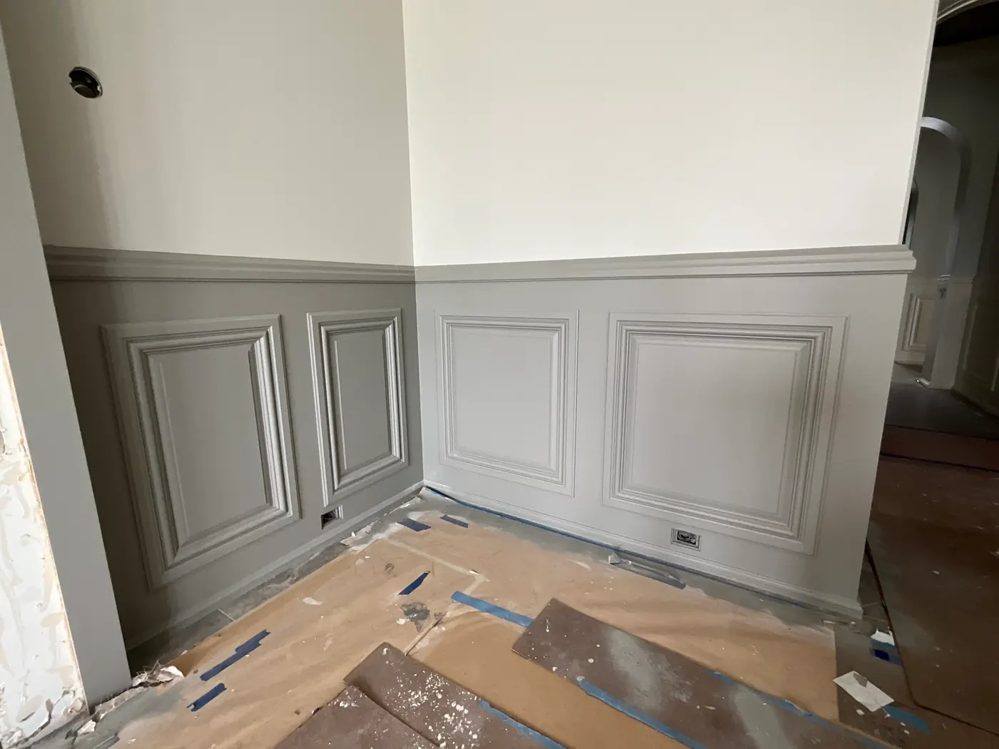 Gray raised-panel wainscoting and chair rail beneath cream-painted upper walls, with the floor covered in protective paper and blue tape.