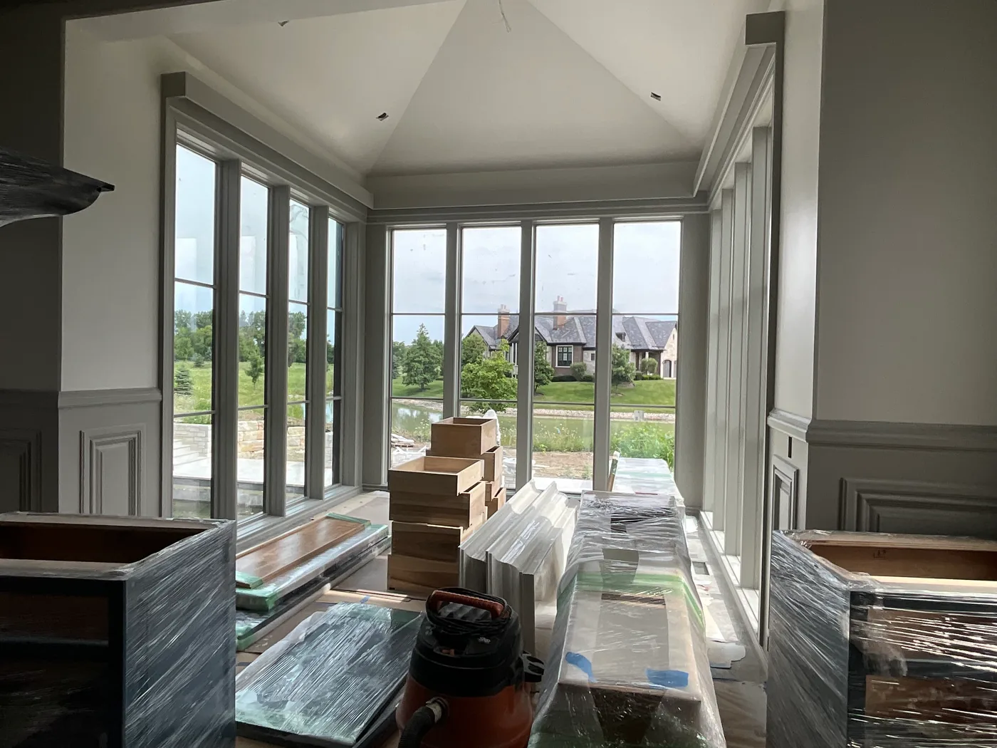 A room with a vaulted ceiling and a bay of tall windows in painted trim looking out over a lawn and pond, with cabinetry and materials wrapped in plastic on the covered floor.